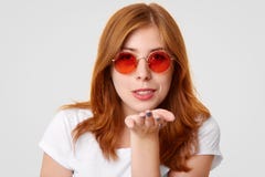 Headshot kokietka imbirowi żeńscy ciosy całuje przy kamerą, wyraża miłości chłopak, jest ubranym modnych round czerwonych okulary obraz stock Headshot kokietka imbirowi żeńscy ciosy całuje przy kamerą, wyraża miłości chłopak, jest ubranym modnych round czerwonych okulary obraz stock