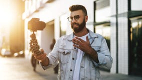 Het gebaarde mannetje hipster blogger in in glazen die zich op stadsstraat bevinden, houdt camera op driepoot en schiet videoblog royalty-vrije stock afbeelding Het gebaarde mannetje hipster blogger in in glazen die zich op stadsstraat bevinden, houdt camera op driepoot en schiet videoblog royalty-vrije stock afbeelding