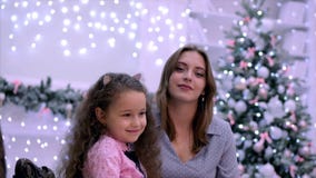 Het jonge aantrekkelijke moeder en dochterzitting koesteren die de camera bekijken die tegen de achtergrond van Kerstmis glimlach stock videobeelden Het jonge aantrekkelijke moeder en dochterzitting koesteren die de camera bekijken die tegen de achtergrond van Kerstmis glimlach stock videobeelden