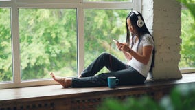 Het leuke Aziatische meisjesbrunette zit op venstervensterbank, luistert aan muziek in hoofdtelefoons en gebruikt smartphone het stock videobeelden Het leuke Aziatische meisjesbrunette zit op venstervensterbank, luistert aan muziek in hoofdtelefoons en gebruikt smartphone het stock videobeelden