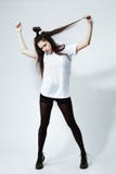 Het modieuze jonge donker-haired meisje met een grappig kapsel gekleed in witte t-shirt, zwarte legging en schoenen stelt op stock afbeelding Het modieuze jonge donker-haired meisje met een grappig kapsel gekleed in witte t-shirt, zwarte legging en schoenen stelt op stock afbeelding