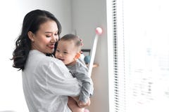 Het mooie jonge mamma speelt met haar leuke baby en glimlacht terwijl thuis status dichtbij het venster royalty-vrije stock foto's Het mooie jonge mamma speelt met haar leuke baby en glimlacht terwijl thuis status dichtbij het venster royalty-vrije stock foto's