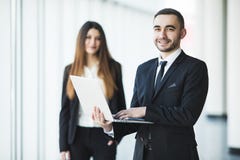 Hombre de negocios joven y confiado con el ordenador portátil que se coloca delante de mujer de negocios imagen de archivo Hombre de negocios joven y confiado con el ordenador portátil que se coloca delante de mujer de negocios imagen de archivo