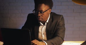 Hombre de negocios negro joven con el ordenador port?til y los papeles que trabajan en la oficina de la noche ?l firma los docume metrajes Hombre de negocios negro joven con el ordenador port?til y los papeles que trabajan en la oficina de la noche ?l firma los docume metrajes