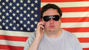 Hombre Gordo En Gafas Sol Que Habla En El Teléfono En El Fondo De Una Bandera De Los E.E.U.U. Almacen De Vídeo de indicador, sunglasses: 146090899