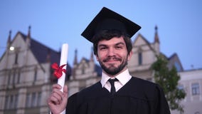 Hombre graduado joven feliz caucásico que presenta a la cámara y que muestra su diploma delante de la universidad metrajes Hombre graduado joven feliz caucásico que presenta a la cámara y que muestra su diploma delante de la universidad metrajes