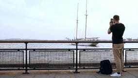 Hombre hermoso joven con la mochila que se coloca en el embarcadero y que toma la foto del barco de motor en el smartphone metrajes Hombre hermoso joven con la mochila que se coloca en el embarcadero y que toma la foto del barco de motor en el smartphone metrajes