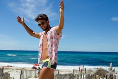 Hombre joven atractivo atractivo en traje de baño y gafas de sol que presentan en la playa con los brazos aumentados en la isla d fotografía de archivo Hombre joven atractivo atractivo en traje de baño y gafas de sol que presentan en la playa con los brazos aumentados en la isla d fotografía de archivo