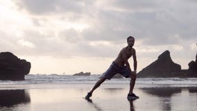Hombre joven en los pantalones cortos y las zapatillas de deporte que hacen estirando ejercicios en una playa con las rocas tempr almacen de metraje de vídeo Hombre joven en los pantalones cortos y las zapatillas de deporte que hacen estirando ejercicios en una playa con las rocas tempr almacen de metraje de vídeo
