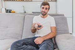 Hombre joven hilarante hermoso que sostiene un teledirigido Durante esto, la TV está mirando mientras que se sienta en el sofá en imagenes de archivo Hombre joven hilarante hermoso que sostiene un teledirigido Durante esto, la TV está mirando mientras que se sienta en el sofá en imagenes de archivo