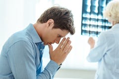 Hombre joven que ruega mientras que el doctor está examinando su fotografía de la radiografía imagenes de archivo Hombre joven que ruega mientras que el doctor está examinando su fotografía de la radiografía imagenes de archivo