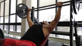 Hombre muscular joven en la prensa de banco Trenes del individuo en el gimnasio con un barbell Hombre muscular joven en la prensa de banco Trenes del individuo en el gimnasio con un barbell