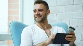 Hombre sonriente joven que hace compras en línea usando la tableta digital que se sienta en el balcón en casa foto de archivo libre de regalías Hombre sonriente joven que hace compras en línea usando la tableta digital que se sienta en el balcón en casa foto de archivo libre de regalías