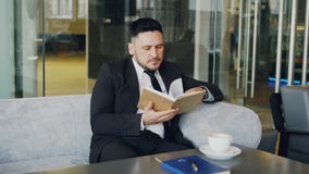 Homem de negócios caucasiano esperto na roupa formal que lê o livro de texto com e que bebe o café no café elegante na pausa para video estoque Homem de negócios caucasiano esperto na roupa formal que lê o livro de texto com e que bebe o café no café elegante na pausa para video estoque