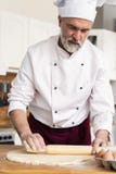 Homem que faz a padaria na cozinha e que rola a massa na placa de corte de madeira coberta com a farinha foto de stock Homem que faz a padaria na cozinha e que rola a massa na placa de corte de madeira coberta com a farinha foto de stock