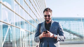 Homme barbu élégant dans des lunettes de soleil utilisant le dispositif tout en passant par le terminal d'aéroport, sourires au t Homme barbu élégant dans des lunettes de soleil utilisant le dispositif tout en passant par le terminal d'aéroport, sourires au t