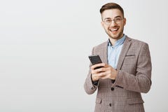 Homme d'affaires avec plaisir bel ambitieux dans les verres et la veste élégante tenant le smartphone regardant fixement l'appare photos stock Homme d'affaires avec plaisir bel ambitieux dans les verres et la veste élégante tenant le smartphone regardant fixement l'appare photos stock
