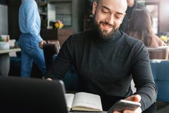 Homme d'affaires barbu travaillant sur l'ordinateur portable tout en se reposant en café, utilisant le smartphone, faisant des en image libre de droits Homme d'affaires barbu travaillant sur l'ordinateur portable tout en se reposant en café, utilisant le smartphone, faisant des en image libre de droits
