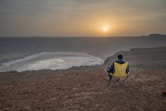 Homme dans une chaise campante à un cratère vulcanic pendant le cratère d'Al Wahbah de lever de soleil en Arabie Saoudite photographie stock libre de droits Homme dans une chaise campante à un cratère vulcanic pendant le cratère d'Al Wahbah de lever de soleil en Arabie Saoudite photographie stock libre de droits
