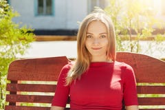 Horizontaal portret van blond vrij jong meisje met sproeten en blauwe ogen die rode sweaterzitting dragen bij bank in parkadmi Stock Foto's Horizontaal portret van blond vrij jong meisje met sproeten en blauwe ogen die rode sweaterzitting dragen bij bank in parkadmi Stock Foto's