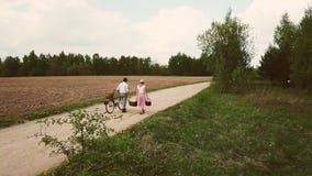 Husband and wife go with a bicycle and baskets go on the road against the background of the field. stock footage Husband and wife go with a bicycle and baskets go on the road against the background of the field. stock footage