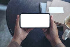 Hände, die einen schwarzen Handy mit leerem Bildschirm horizontal für das Aufpassen mit Kaffeetasse und Notizbüchern auf Tabelle stockfotografie Hände, die einen schwarzen Handy mit leerem Bildschirm horizontal für das Aufpassen mit Kaffeetasse und Notizbüchern auf Tabelle stockfotografie