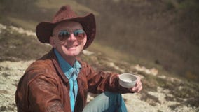 I bergen piskar en man i en cowboyhatt, omslaget, jeans och exponeringsglas Mannen ser ramen och leendena stock video I bergen piskar en man i en cowboyhatt, omslaget, jeans och exponeringsglas Mannen ser ramen och leendena stock video