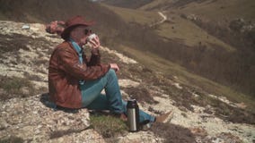 I bergen sitter en man i en cowboyhatt, piskar omslaget, jeans och exponeringsglas E arkivfilmer I bergen sitter en man i en cowboyhatt, piskar omslaget, jeans och exponeringsglas E arkivfilmer