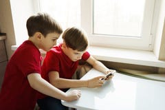I due fratelli stanno sedendo nella cucina e stanno giocando con il telefono immagine stock I due fratelli stanno sedendo nella cucina e stanno giocando con il telefono immagine stock