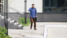 Il giovane uomo bello vestito in camicia casuale blu e pantaloni marroni sta uscendo dalla costruzione che parla sul video d archivio Il giovane uomo bello vestito in camicia casuale blu e pantaloni marroni sta uscendo dalla costruzione che parla sul video d archivio