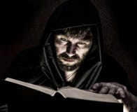 Il negromante lancia gli incantesimi dal libro antico spesso da lume di candela su un fondo scuro fotografia stock Il negromante lancia gli incantesimi dal libro antico spesso da lume di candela su un fondo scuro fotografia stock