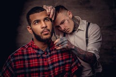 Il parrucchiere tatuato professionista antiquato fa un taglio di capelli ad un cliente afroamericano su buio strutturato fotografia stock Il parrucchiere tatuato professionista antiquato fa un taglio di capelli ad un cliente afroamericano su buio strutturato fotografia stock