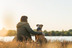 Il proprietario del cane ed il suo animale domestico si siedono alla riva al tramonto immagine stock Il proprietario del cane ed il suo animale domestico si siedono alla riva al tramonto immagine stock