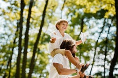 Il ragazzo biondo allegro ed il padre bello vestiti nelle magliette bianche sono nel parco Il ragazzo si siede sulle spalle del p immagine stock Il ragazzo biondo allegro ed il padre bello vestiti nelle magliette bianche sono nel parco Il ragazzo si siede sulle spalle del p immagine stock