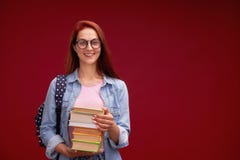 Il ritratto di bella studentessa con uno zaino e di una pila di libri in sue mani sta sorridendo ai precedenti rossi fotografie stock libere da diritti Il ritratto di bella studentessa con uno zaino e di una pila di libri in sue mani sta sorridendo ai precedenti rossi fotografie stock libere da diritti