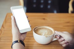 Il ` s della donna passa la tenuta del telefono cellulare bianco con lo schermo in bianco e la tazza di caffè macchiato di latte Fotografia Stock Il ` s della donna passa la tenuta del telefono cellulare bianco con lo schermo in bianco e la tazza di caffè macchiato di latte Fotografia Stock