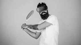 Il tipo con i vetri di VR gioca a tennis con la racchetta e la palla L'uomo con la barba in vetri di VR gioca a tennis, fondo gri fotografia stock libera da diritti Il tipo con i vetri di VR gioca a tennis con la racchetta e la palla L'uomo con la barba in vetri di VR gioca a tennis, fondo gri fotografia stock libera da diritti