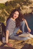 Il viaggiatore con zaino e sacco a pelo della ragazza sta sedendosi sul libro di lettura e della roccia fotografie stock Il viaggiatore con zaino e sacco a pelo della ragazza sta sedendosi sul libro di lettura e della roccia fotografie stock