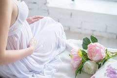 Image en gros plan de femme enceinte dans la robe blanche intéressante touchant son ventre avec des mains et tenant un bouquet de photographie stock libre de droits Image en gros plan de femme enceinte dans la robe blanche intéressante touchant son ventre avec des mains et tenant un bouquet de photographie stock libre de droits