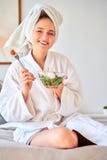 Image of young woman in bathrobe with salad in bowl in her hands sitting on bed.
