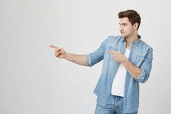 Indoor shot of confident handsome young guy looking and pointing left while standing over gray background. Attractive stock images Indoor shot of confident handsome young guy looking and pointing left while standing over gray background. Attractive stock images