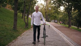 Integral del hombre joven sonriente del caucásico en la camisa blanca que camina con la bicicleta en la calle en ciudad Rueda el almacen de video Integral del hombre joven sonriente del caucásico en la camisa blanca que camina con la bicicleta en la calle en ciudad Rueda el almacen de video