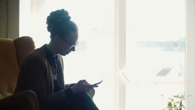 Jeune belle femme s'asseyant dans la chaise jaune près de la fenêtre et à l'aide du smartphone, causant dans l'Internet banque de vidéos Jeune belle femme s'asseyant dans la chaise jaune près de la fenêtre et à l'aide du smartphone, causant dans l'Internet banque de vidéos