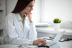 Jeune docteur féminin dactylographiant sur l'ordinateur portable tout en se reposant à la table près de la fenêtre dans le bureau photo stock Jeune docteur féminin dactylographiant sur l'ordinateur portable tout en se reposant à la table près de la fenêtre dans le bureau photo stock