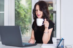 Jeune et mignonne femme d'affaires travaillant sur l'ordinateur portable et tenant la tasse de café images libres de droits Jeune et mignonne femme d'affaires travaillant sur l'ordinateur portable et tenant la tasse de café images libres de droits