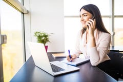 Jeune femme attirante parlant au téléphone portable et souriant tout en se reposant à son lieu de travail dans le bureau et regar Photographie stock