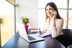 Jeune femme attirante parlant au téléphone portable et souriant tout en se reposant à son lieu de travail dans le bureau et regar Photographie stock