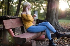 Jeune femme blonde s'asseyant sur un banc en bois en parc, écoutant la musique Photographie stock Jeune femme blonde s'asseyant sur un banc en bois en parc, écoutant la musique Photographie stock