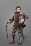 Jeune femme dans le chevalier Armour tenant le casque dans une main et épée sur son épaule au-dessus de fond gris photos stock Jeune femme dans le chevalier Armour tenant le casque dans une main et épée sur son épaule au-dessus de fond gris photos stock