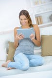 Jeune femme gaie s'asseyant sur le divan avec le comprimé image libre de droits Jeune femme gaie s'asseyant sur le divan avec le comprimé image libre de droits